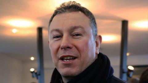 A man with short, dark hair looks directly at the camera. He is in a room with spotlights and two large black pillars behind him. The man is wearing a dark coat and a scarf, draped loosely around his neck. 