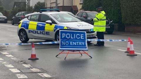 A police car and a police officer by a closed road, with a blue police sign placed on the road. There are red and white traffic cones and tape across the road. Other cars are in the street and a row of houses, to the right. There are also green bushes to the right. 
