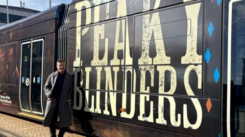 A man in a long grey coat stood next to a tram with the words Peaky Blinders emblazoned on the sides in cream lettering