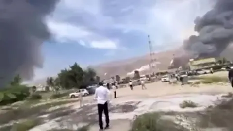 Two large plumes of smoke while people stand in a car park and look at the scene - the image is low quality and hard to make out 
