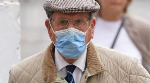 A man wearing a white shirt, navy tie and cream coat. He is also wearing a blue facemask.