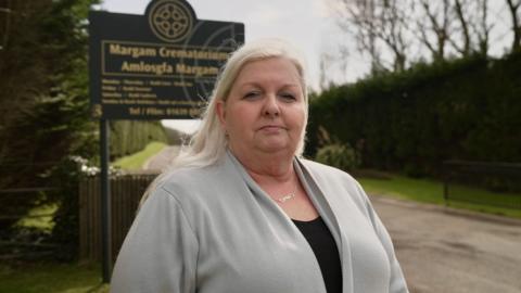 Katherine James - menyw gwallt melyn hir mewn top du a chardigan lwys - yn sefyll wrth fynedfa Amlosgfa Margam