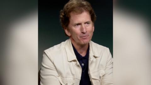 A man in a dark top with a cream jacket sits down