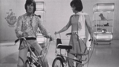 Black and white image of Peter Purves and Valerie Singleton on chopper bikes in the Blue Peter studio.  In the background are a couple pieces of scenary and a Blue Peter ship logo hanging from the ceiling.  Peter is astride his bike and looking directly down the lens.  Valerie is standing beside her bike and looking at Peter.