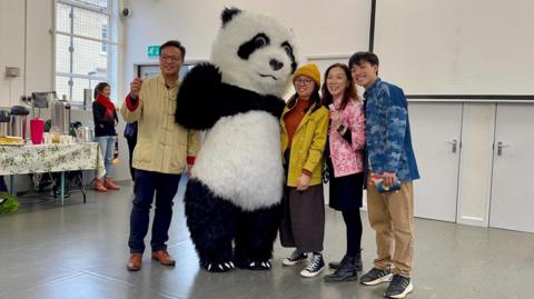 A large panda mascot stood waving in among a family of people from Hong Kong. They are smiling and looking at the camera. They are standing in a building with white walls and doors.
