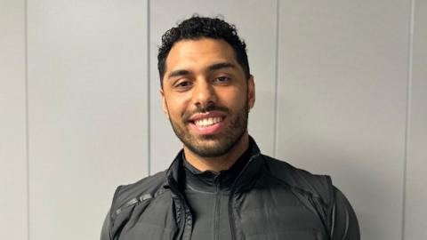 A picture of Leon Greenwood in the BBC Radio Berkshire offices. He is wearing a black zip-up top with a black gilet. He has short dark hair and is looking at the camera and smiling.