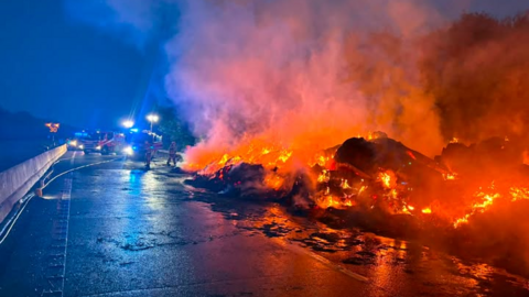 A huge pile of material on fire on a motorway at night. Firefighters and fire engines with blue lights can be seen in the background.