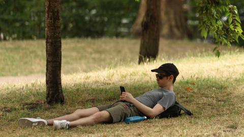 A man wearing a baseball cap and sunglasses using a mobile phone while lying down on yellow dry-looking grass, under trees