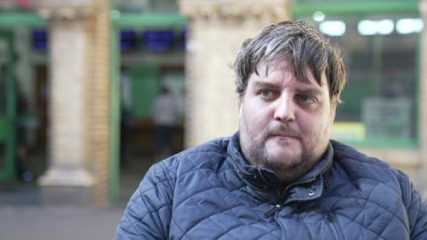 Nathan Rollinson is seen with short brown hair and a beard wearing a navy quilted jacket, pictured in front of a brick wall and green doorways.