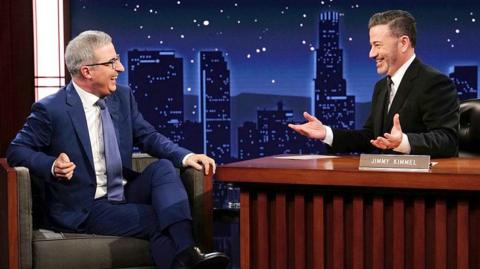 John Oliver sits with Jimmy Kimmel on set during a talk show, they are both smiling and sitting against a night sky cityscape backdrop