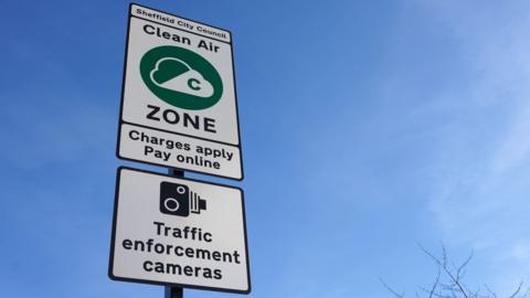 A clean air zone road sign. There is a large green circle with the outline of a cloud half coloured white. Either side of the symbol reads "Clean air" then "ZONE". Two smaller signs say "charges apply, pay online" and "traffic enforcement cameras".