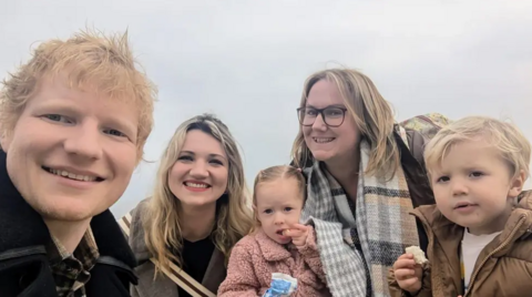 Ed Sheeran poses for a selfie with Amanda Jane, her friend, and their children. Ed has ginger hair and is smiling into the camera. Amanda is wearing glasses and a scarf while her female friend has blonde hair. The two children pictured are each holding a piece of food and staring into the camera.
