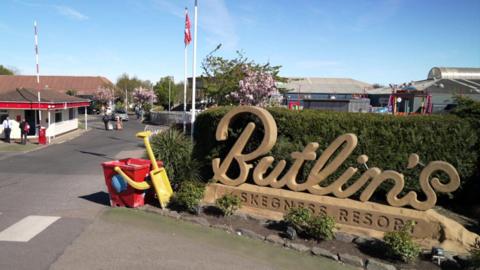 The entrance to a Butlin's resort, dominated by a large sand-coloured sign reading "Butlin's" in script and "Skegness Resort" in capital letters. Behind the sign is a tall green hedge and next to it a giant red bucket and yellow spade. In the background, a security hut, red and white raised barrier and resort buildings can be seen under a blue sky.