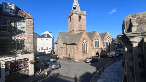 Town church with blue skies in the background. Cars parked on a road. 