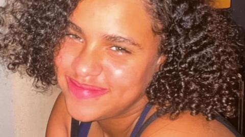 Emily, smiling at the camera. She has brown curly hair and wears a vest top. 