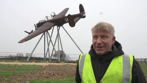 Charlie White stood in front of a life-size steel sculpture of a Lancaster Bomber. He has short blond hair and is wearing a black zip-up, hooded coat and a yellow hi-vis vest. The sky is overcast. 