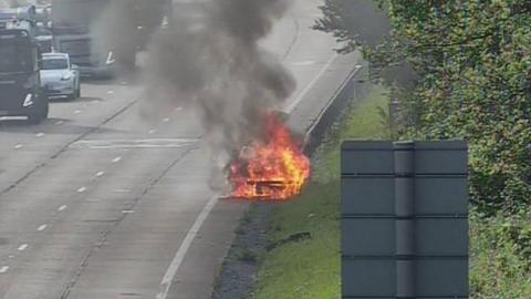 A vehicle fully engulfed in flames on the side of the road. There is a grassy bank and trees on the right.