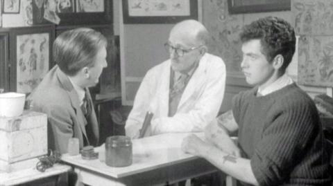 The reporter, a tattoo artist wearing a lab coat, and a man with tattoos on his arms sit around a small table