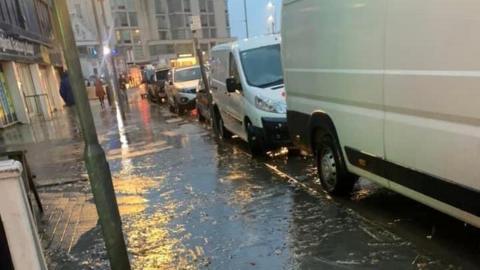 A pavement covered in floodwater on a wet and dark evening. To the right of the image are several parked cars and vans. The water is flowing off the pavement. There are items of debris in the water that appear to be raw sewage.