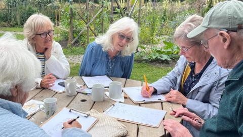 Four women and one man sitting around a round wooden table in a garden. There are plants and flowers in the background.  The people all have clip boards and paper with drawings on on the table in front of them and two of the ladies are drawing with pencils, while the teacher, a woman with white shoulder length hair, glasses and a denim shirt looks on.  There are also four empty mugs on the table. 