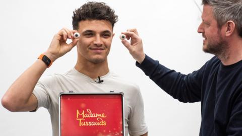 F1 driver Lando Norris in a t shirt, holding up fake eyes from a box labelled Madame Tussauds.