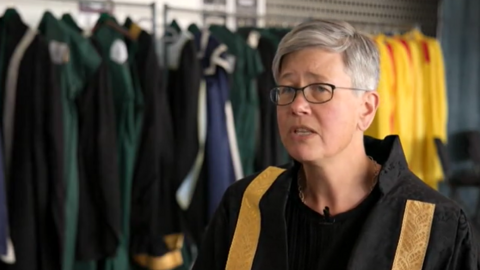 A woman with short grey hair and glasses wearing a black robe with gold trim. In the background is a clothes rail with green and yellow robes hung up