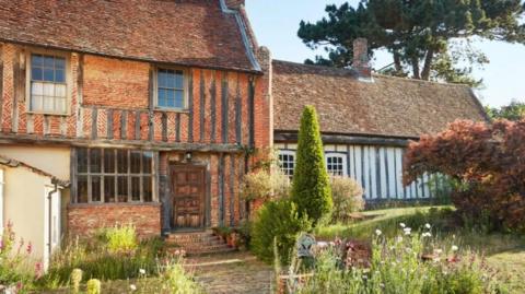 A Tudor home and a garden in front of it. The home is made of red brick and wooden beams. 