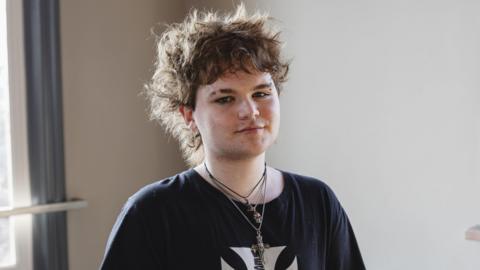 A young man in a black t-shirt, wearing three necklaces . He is looking at the camera in front of a plain background.