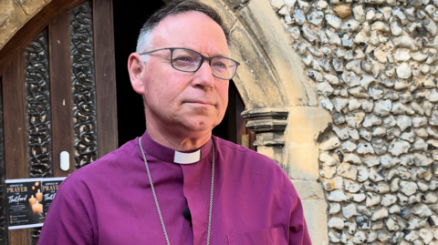 A bishop, dressed in a purple shirt and clerical collar, is standing outside a stone church.