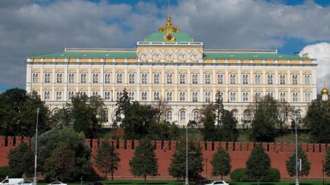 Picture of Kremlin Palace during the day. There is a blue sky and clouds. In front of the palace is a red brick wall and trees lined in front of it and behind it