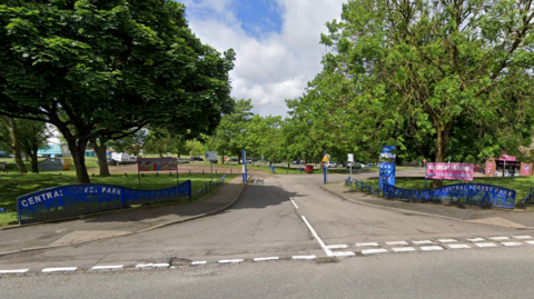 The entrance to a car park, which has blue signage indicating it is Central Forest Park. There are white dashed give way markings at the junction with the road, and the car park is surrounded by trees.