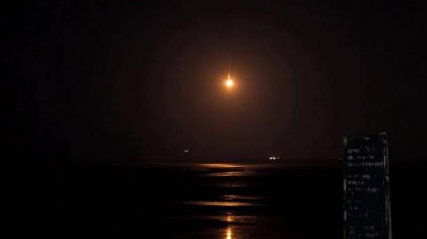 Image shows a near pitch black beach with the distant light in the sky, potentially from a police helicopter or a flare.