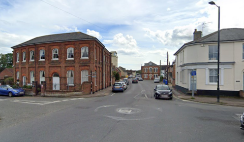 A street view of a small mini-roundabout intersection in a UK town, featuring a prominent two-story red brick historical building on the left corner and cars parked along the road stretching into the background under a partly cloudy sky.