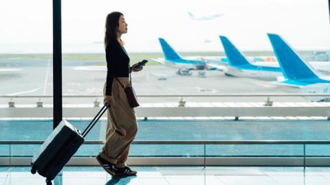 A woman is walking through an airport pulling along a suitcase and holding her phone in her hand. Three planes are parked behind her on the tarmac.