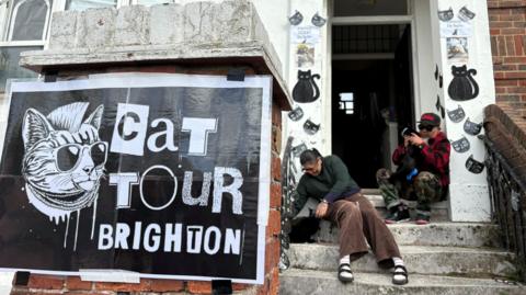 Two cat owners and volunteers for Brighton Cat Tour sit on concrete steps outside their flat. On the red brick column in front of the steps and doorway sits a punk-themed poster reading "Cat Tour Brighton", depicting a cat with a mohawk, sunglasses and chains sticking its tongue out. The owners sit and pet two black cats, with black cat-themed decorations adorning the front entryway to their home for passing visitors.