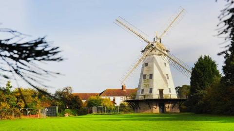 A 19th Century windmill sits among semi-rural grassland. It is made of light coloured wood and has four sails. 