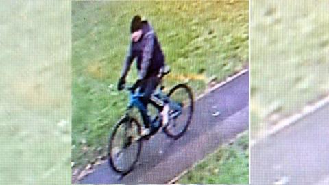 A CCTV image of a man on a bike, wearing a tracksuit and a black hat, cycling along a path in a park