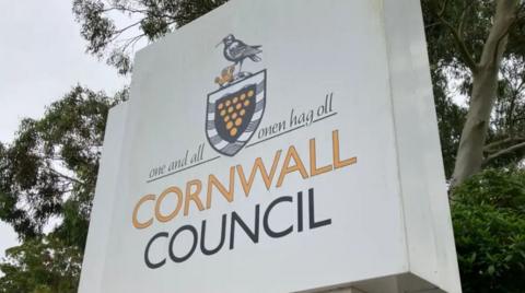 Cornwall Council sign - white background, yellow and black writing with the Cornish crest. Trees behind and grey sky.