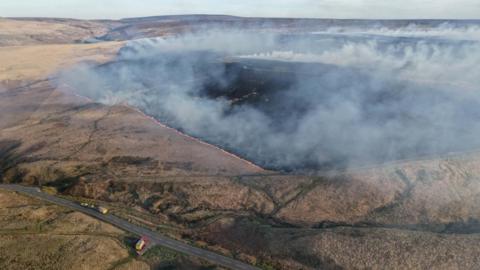 An aerial view showing a large expanse of moorland with smoke rising from the ground and orange flames around a section of blackened land
