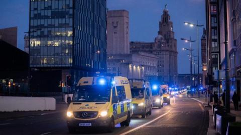 A convoy of yellow police vans in front of the Liver Building