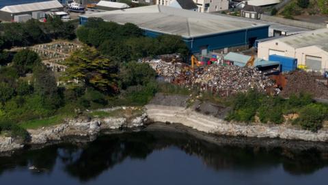 A drone shot showing the proximity between the Guernsey Scrap Metal, the St Sampson's Parish church and Longue Hougue reservoir. Scrap is falling from a large mountain of waste into the quarry. 