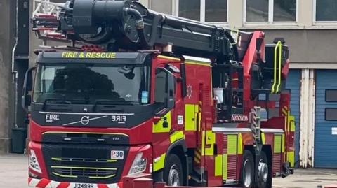 A red and yellow fire engine, facing the camera. Behind it is a grey block of flats with a blue door. There are windows on the second floor. 