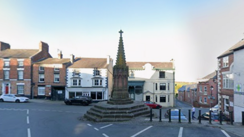 A village cross, a stone monument with an elaborate cone on top of a set of steps on a hexagonal platform. There is a road either side and car parking spaces next to the monument. There are buildings in the background of the image with parked cars outside them.