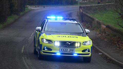 A Cheshire Police car with its blue emergency lights on driving down a road. There is a pavement to the side with leaves on. 