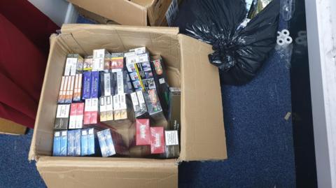 An open brown cardboard box containing multiple packets of cigarettes. Also a black bin liner in the top left corner and till rolls on the floor.