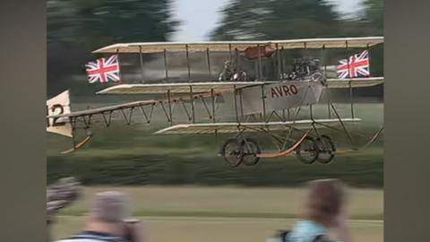 An aircraft taking off as part of the Shuttleworth air show. There are flags on the plane and the word AVRO is on the side of it in red lettering.