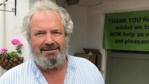 David Beavan, who has grey hair and a beard and is wearing a collarless shirt, smiles as he stands in front of a building