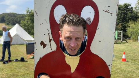 Matt Larcombe. A man is stuck in stocks, namely a board chick, with his head poking through. He has wet brown hair and is smiling. Behind him is a green field. 