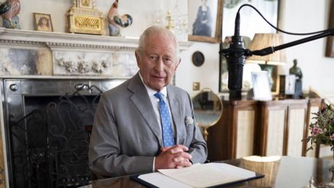 King Charles III, wearing a blue tie and grey suit, looks directly down the camera lens, a radio microphone is hangs about him ready to give his address