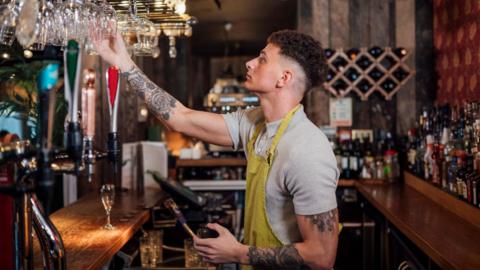 A bar staff member wearing a grey t-shirt and a yellow apron stands behind the bar holding a glass in his hand.